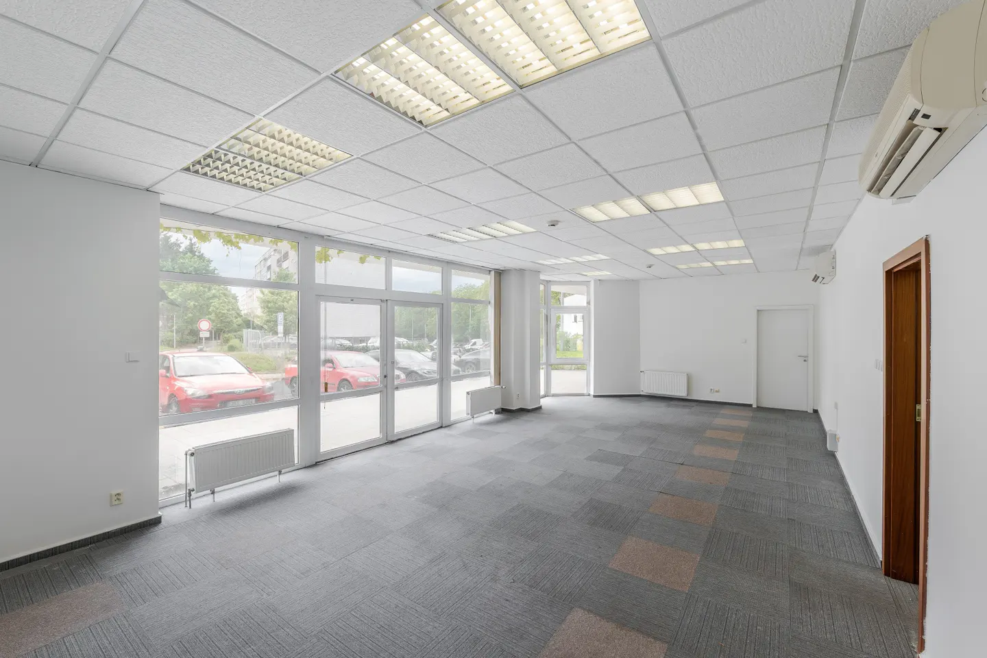 Bright, empty office space with gray carpet, white walls, and large windows showing red cars outside.
