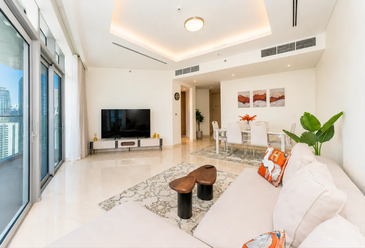 Bright, modern apartment living room with a white sofa, TV, dining table, and city views from large windows.