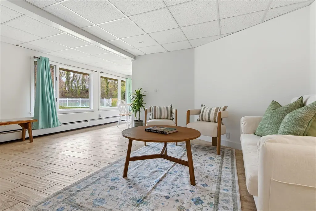 Bright living room with white walls, wood floors, and a blue patterned rug. A round wood table sits in front of two chairs and a white sofa. Large windows let in natural light.