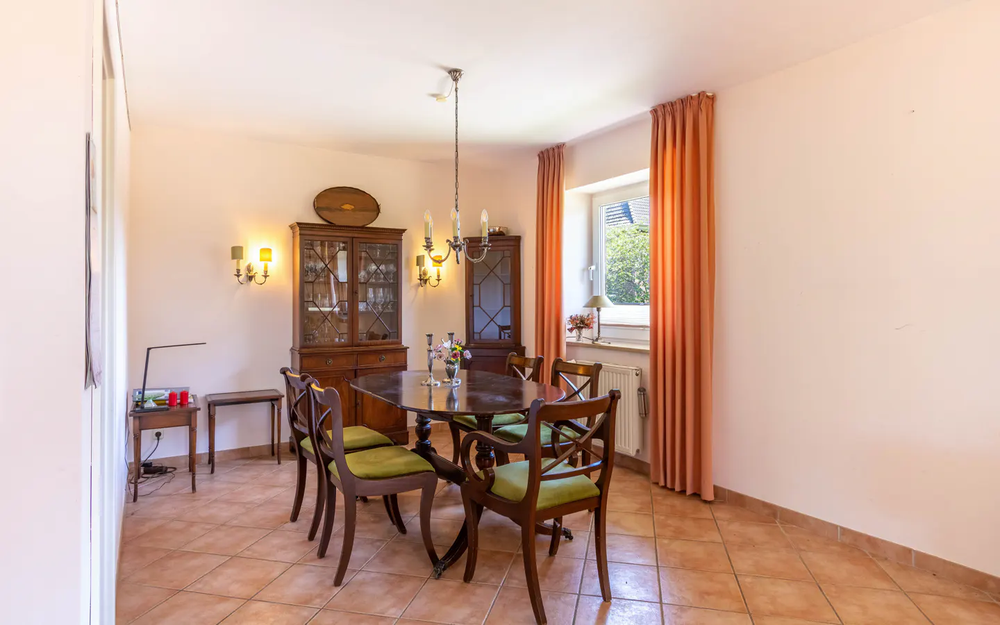 Dining room with a dark wood table, green chairs, a matching hutch, and orange curtains by a window on a tiled floor.