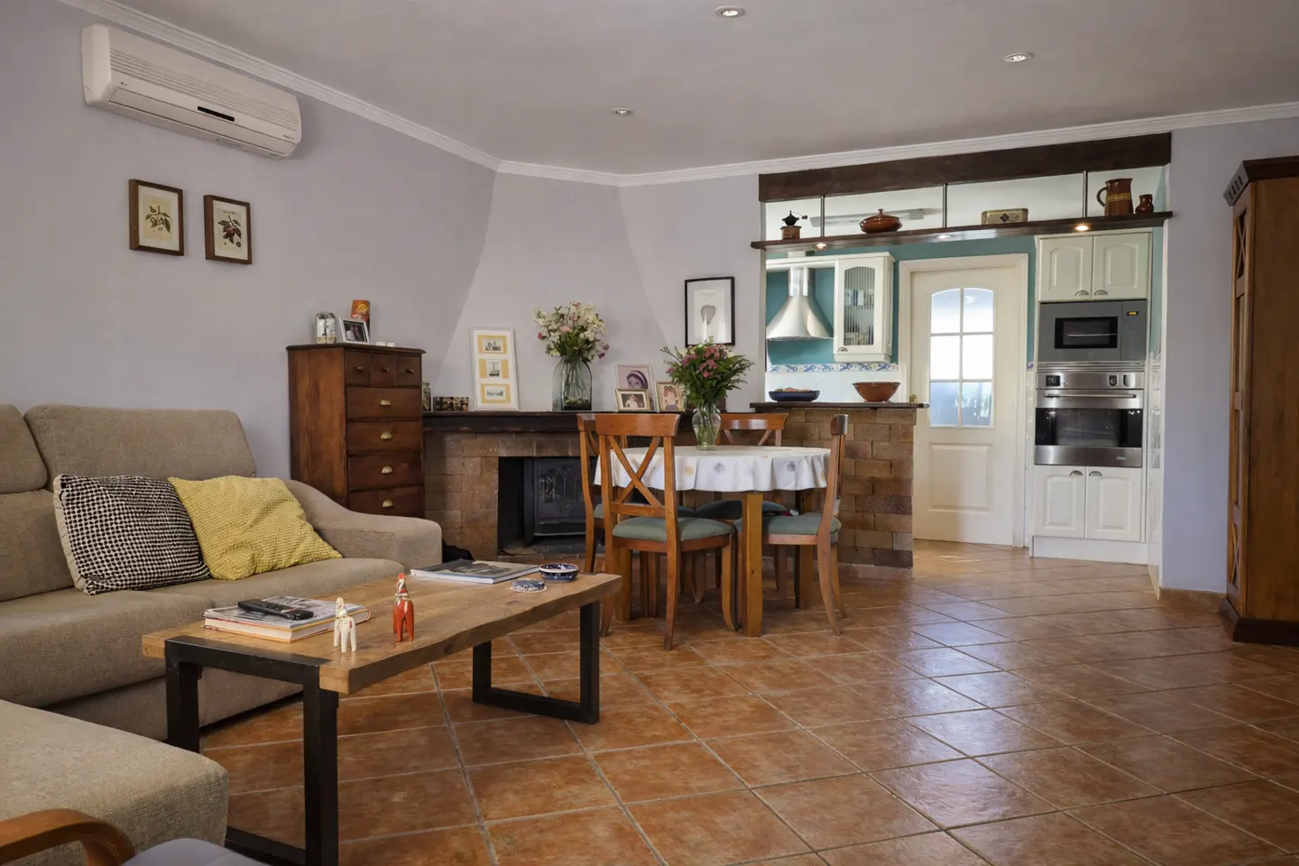 Living room with a beige sofa, wooden table, and dining set. A fireplace and kitchen entrance are visible in the background.