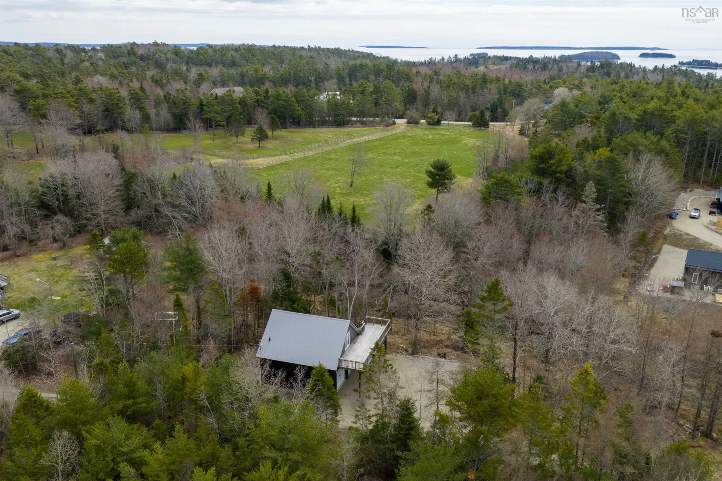 Aerial view of a dark house with a gray roof surrounded by trees, a green field, and a body of water in the distance.