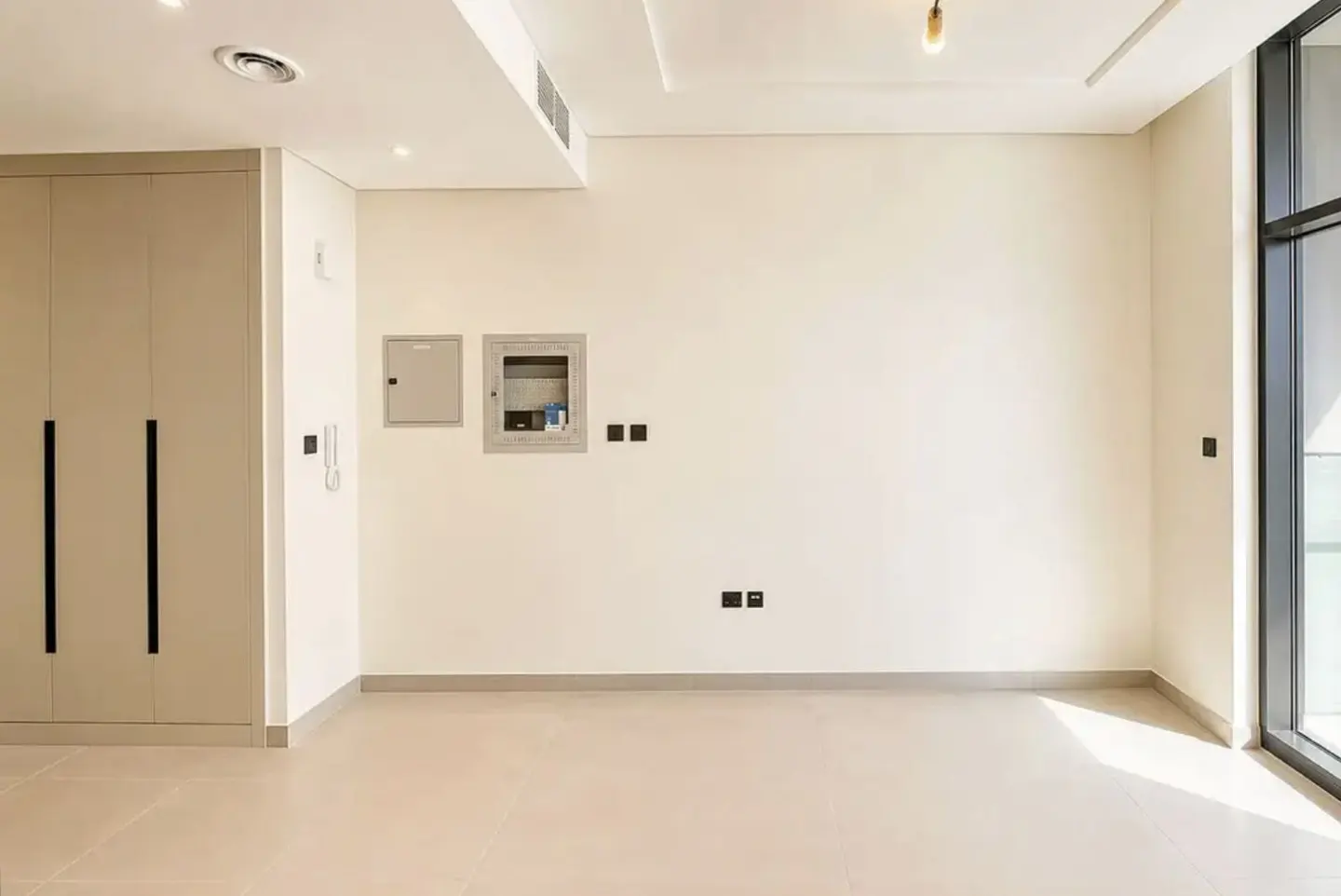 An empty room with beige walls and tile floors, featuring a closet with black handles and a large window.