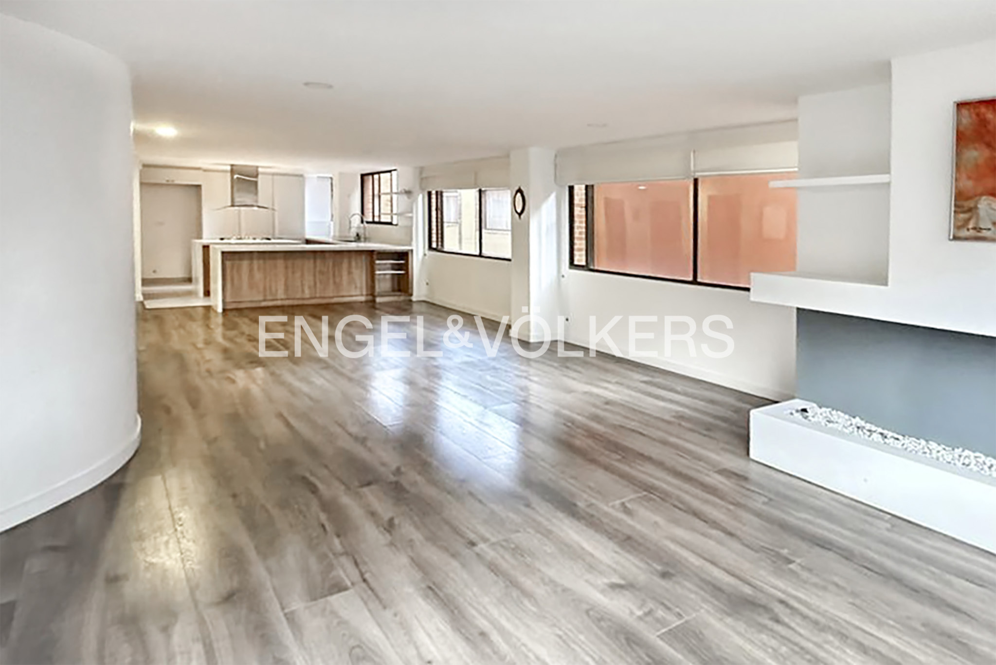Open-concept living space with wood floors, white walls, and a kitchen island. Large windows provide natural light.