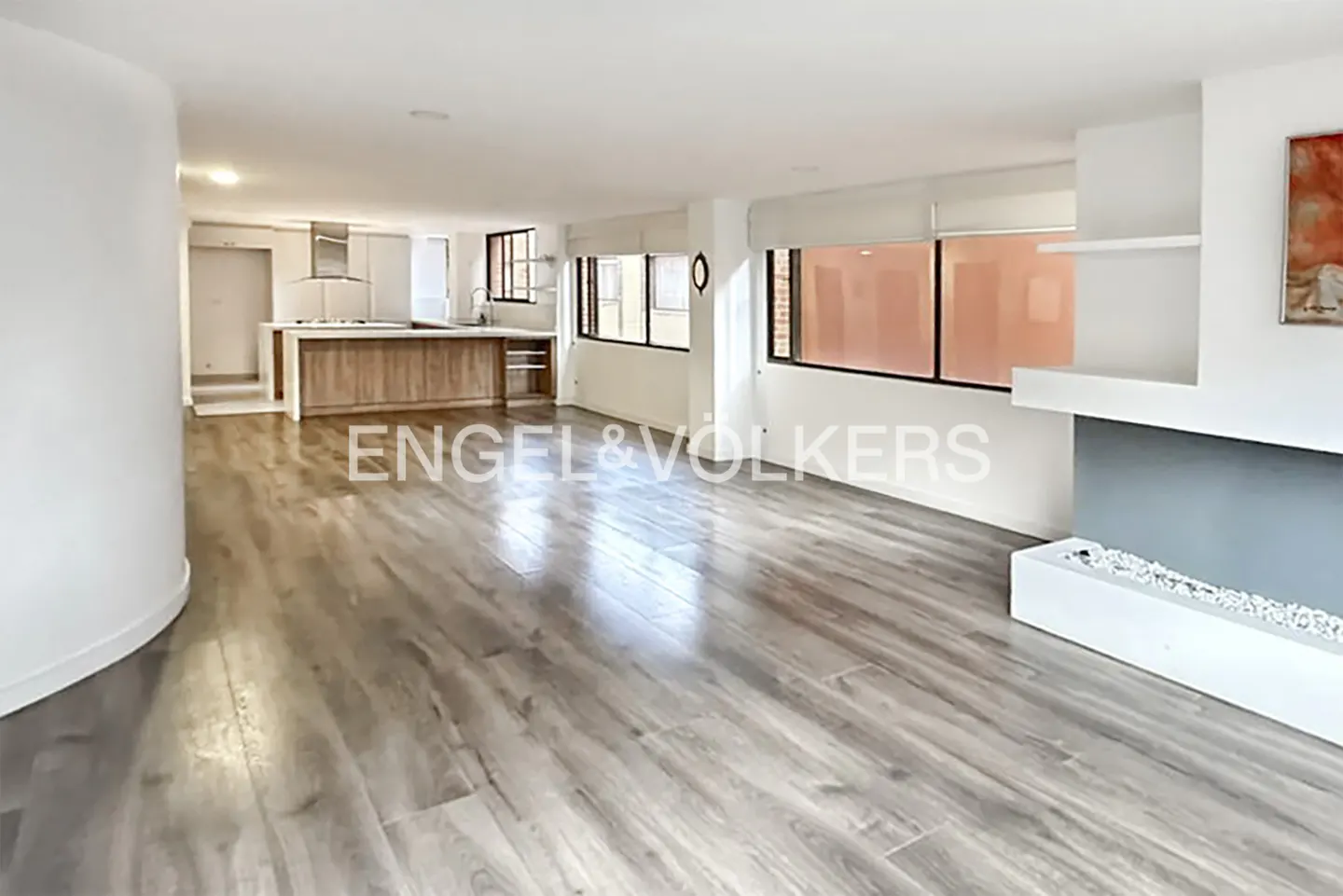 Open-concept living space with wood floors, white walls, and a kitchen island. Large windows provide natural light.