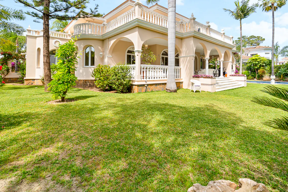 Exterior view of a beige two-story house with arched porch, white railings, and green lawn.