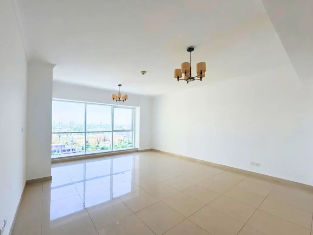 Bright, empty room with beige tile floor, white walls, and large window showing city view. Two modern chandeliers hang from the ceiling.