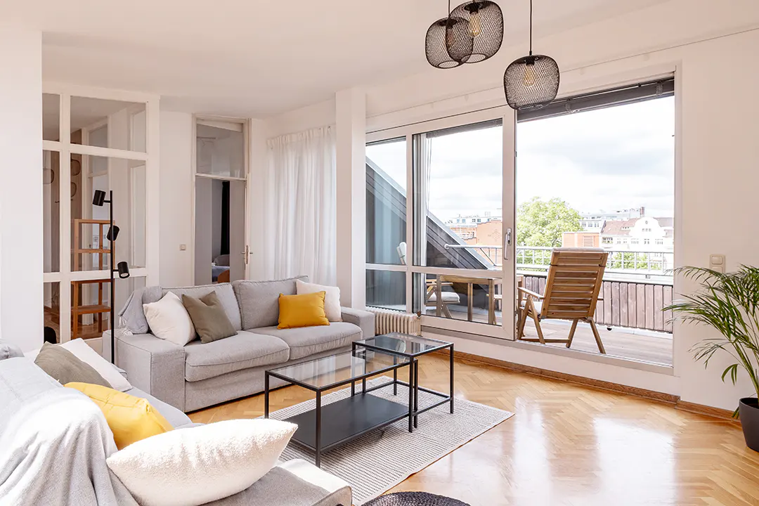 Bright living room with gray sofas, yellow pillows, and a glass coffee table. Balcony with wooden furniture visible through large windows.