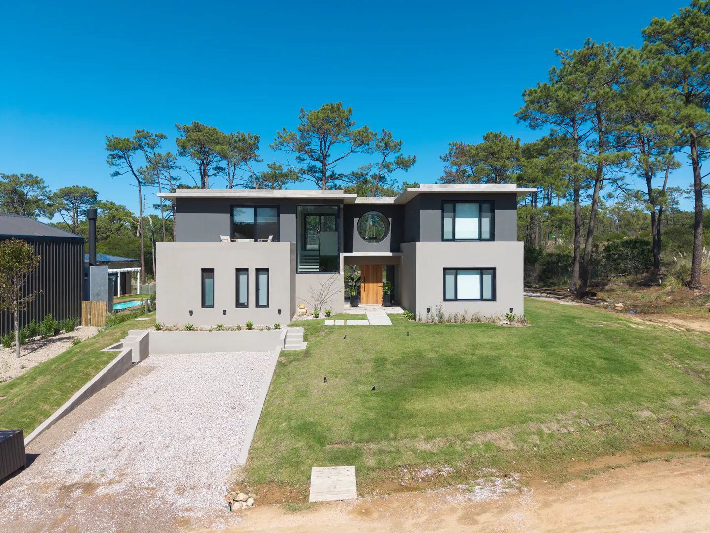 Two-story modern house with gray and beige exterior, a round window, and a gravel driveway on a green lawn.