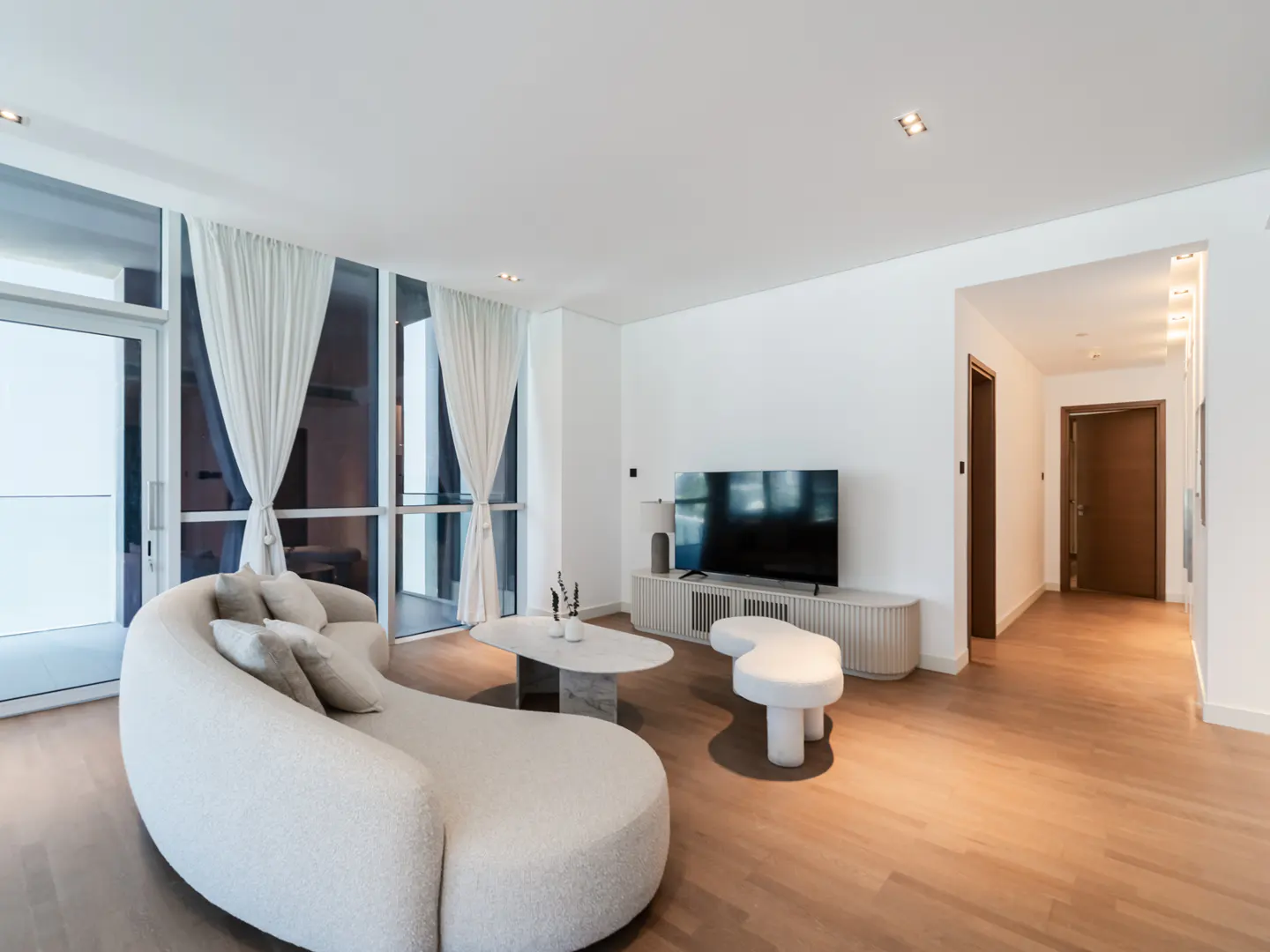 Bright living room with a curved white sofa, marble table, and TV. Large windows with white curtains let in natural light. Wooden floors add warmth.