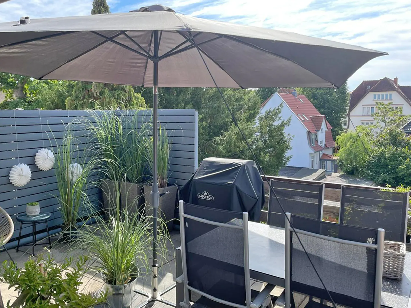 Outdoor patio with gray umbrella, grill, table, chairs, and potted plants. Gray fence with white decorations. Houses and trees in the background.