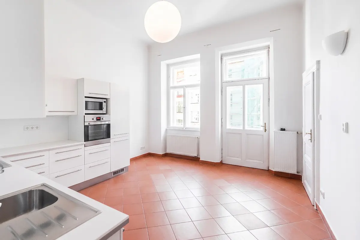 Bright, empty kitchen with white cabinets, stainless steel sink, and terracotta tile floor. Large windows and a white door let in natural light.