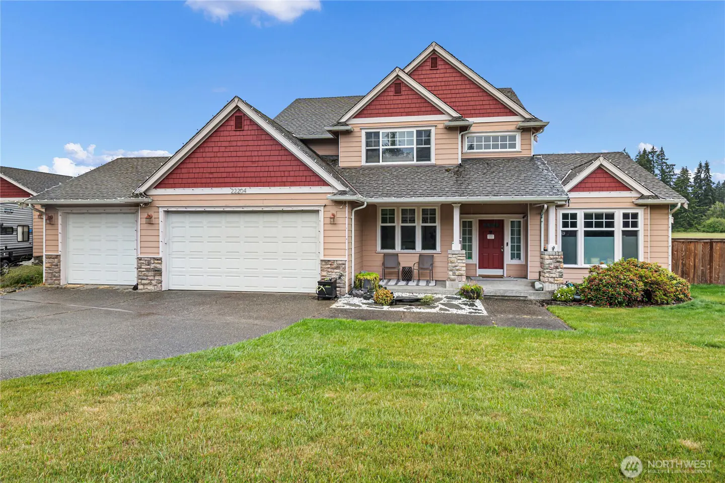 Two-story suburban home with tan siding, red shingle accents, a three-car garage, and a front porch on a grassy lawn.