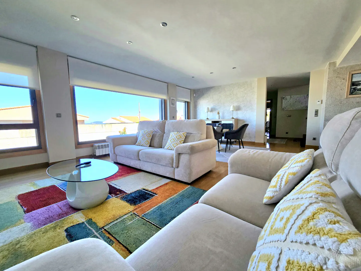 Bright living room with two beige sofas, a glass coffee table, and a colorful geometric area rug under large windows.