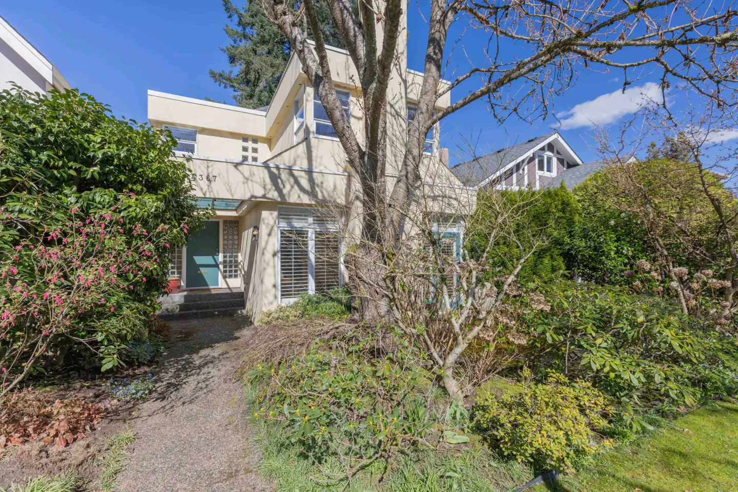 Two-story beige house with a teal door and white-framed windows, surrounded by lush green bushes and trees.