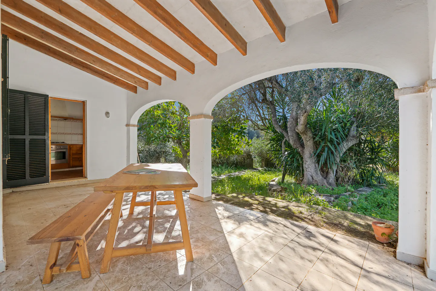 Covered patio with wooden table and bench, white columns, and arched openings to a green garden. Interior kitchen visible through open door.