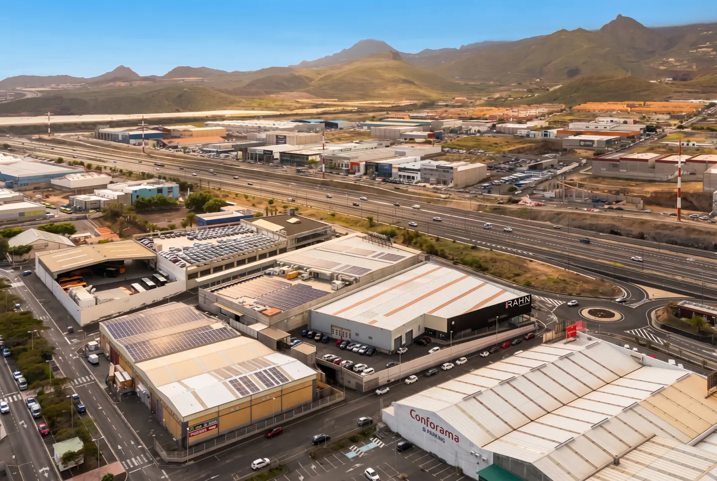 Aerial view of a commercial area with warehouses, parking lots, and a highway, set against a backdrop of mountains under a blue sky.