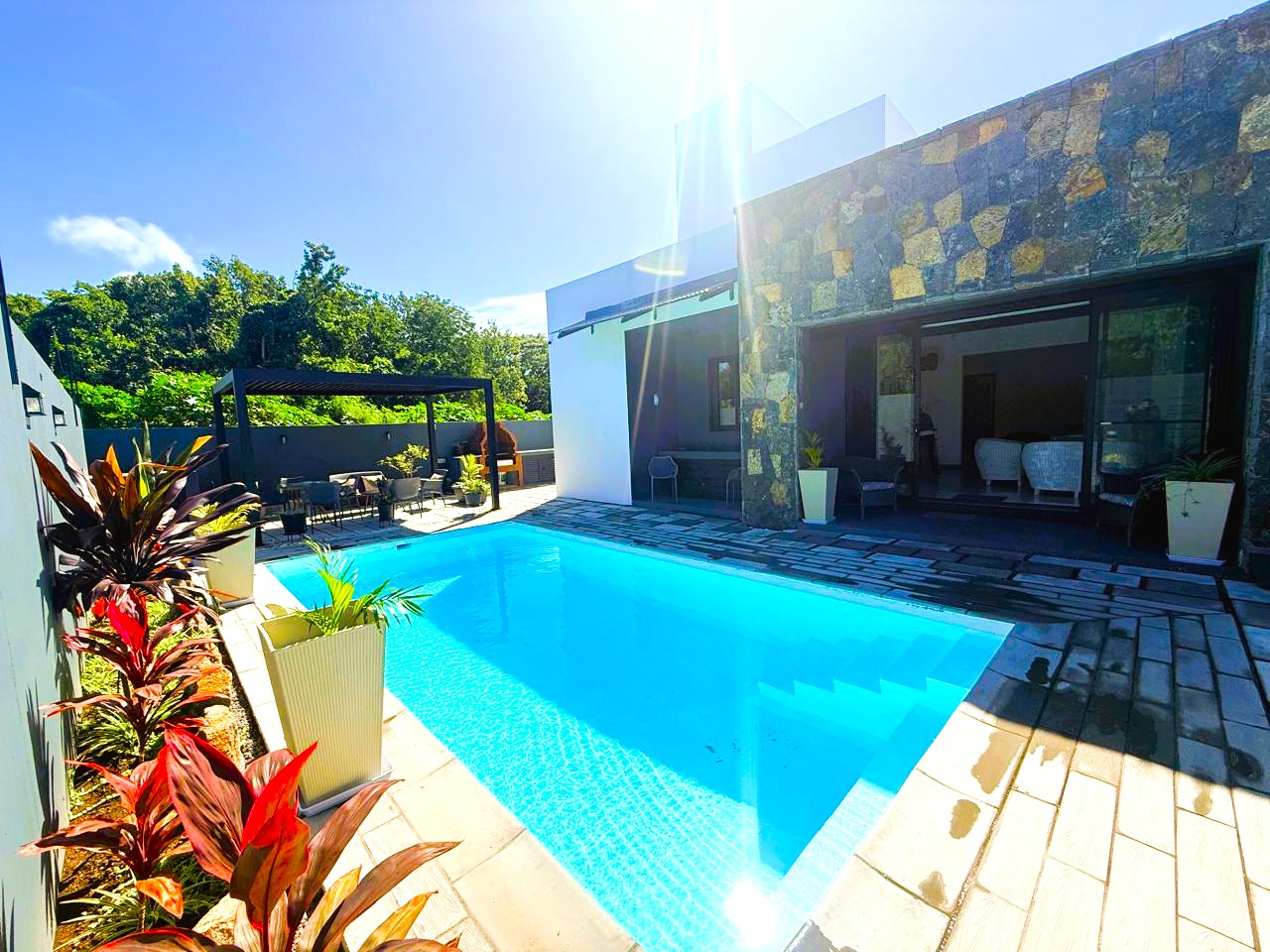 Bright blue swimming pool on a stone patio next to a modern home with a stone facade and a shaded outdoor seating area.