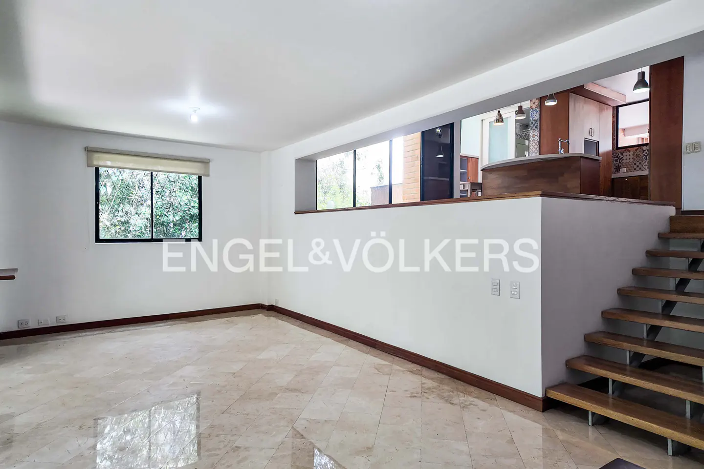 Bright, empty living room with marble floors, white walls, and a window. Stairs lead to a kitchen area with wood accents. Engel & Völkers logo on the wall.