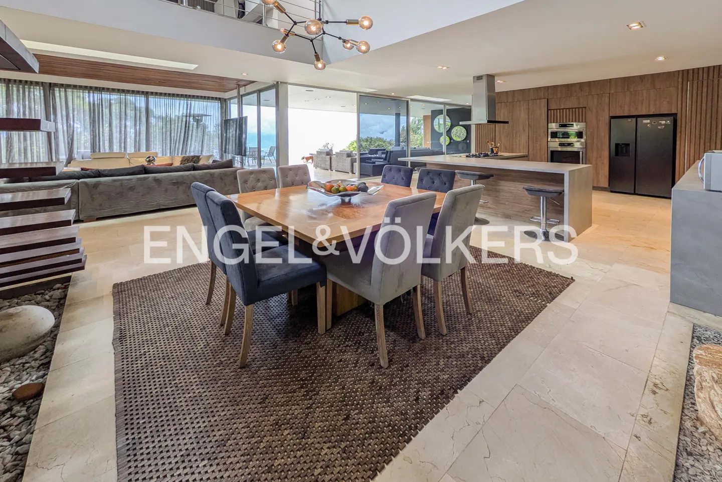 Open-concept living space with a wood dining table, gray chairs, and a brown rug. Kitchen island and ocean view in the background.
