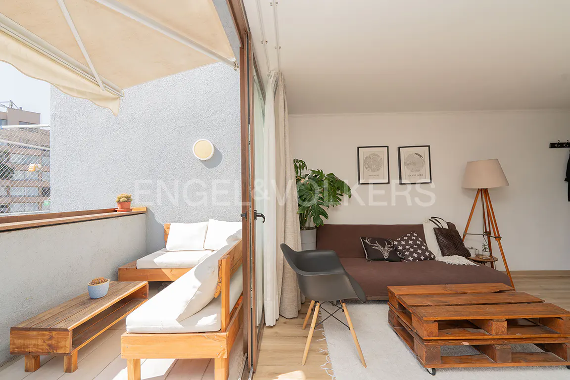 Bright living space with a balcony. The balcony has wooden furniture with white cushions. Inside, a brown sofa, pallet coffee table, and a floor lamp.