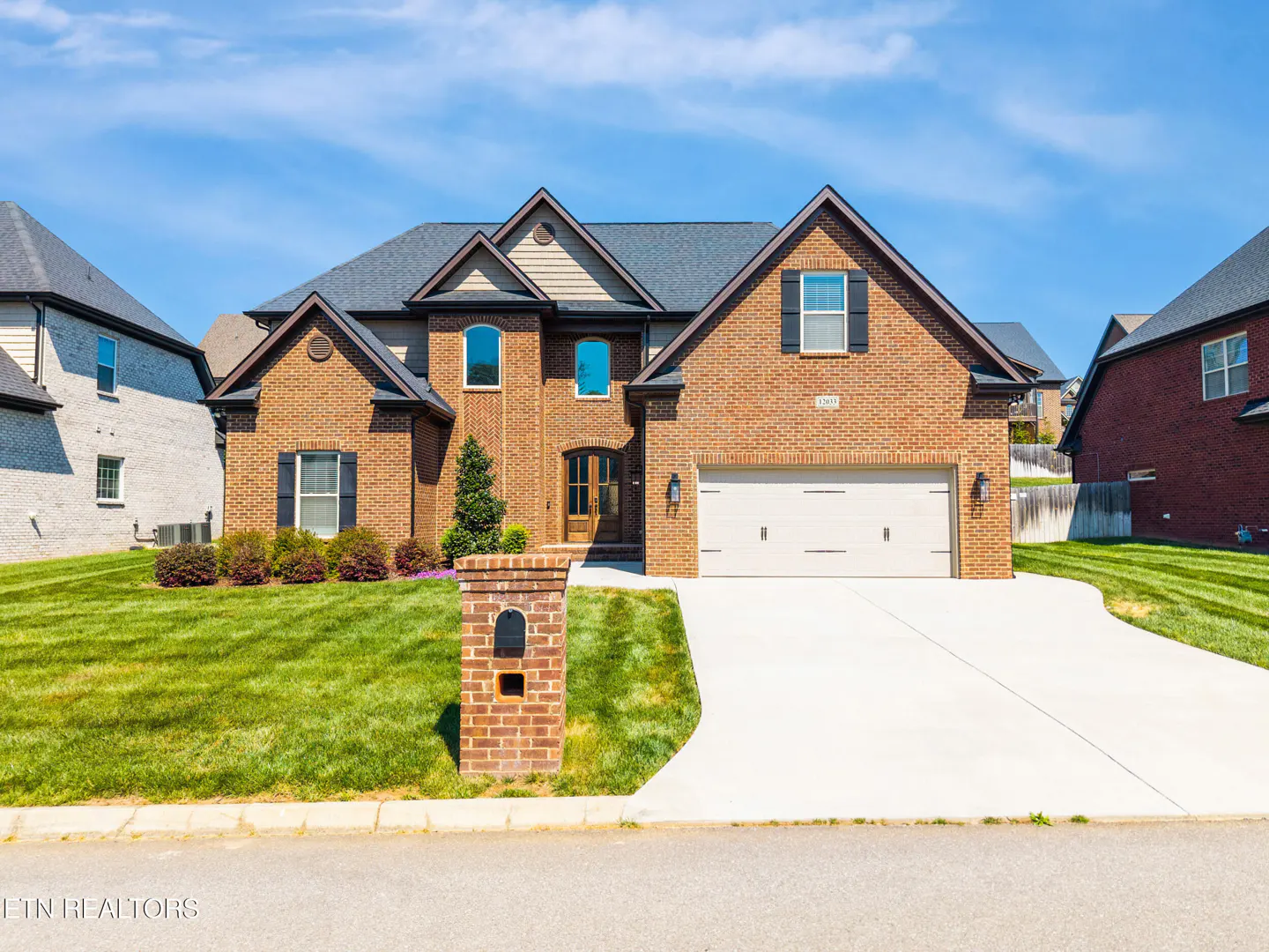 Two-story brick house with a gray roof, white garage door, and green lawn under a blue sky.