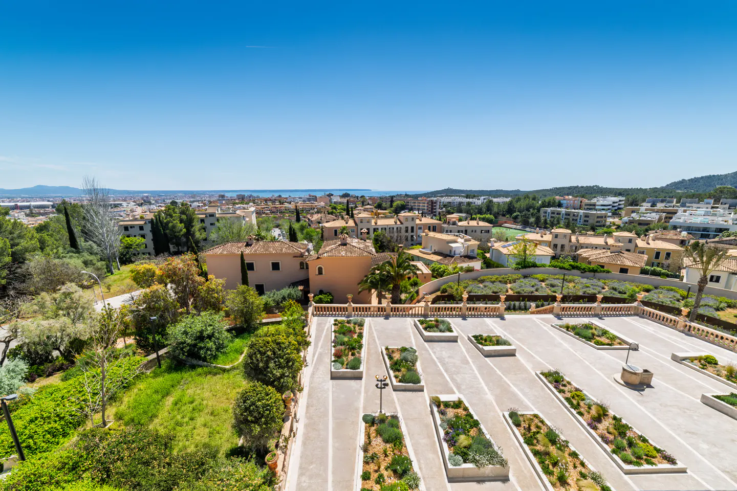 Aerial view of a Mediterranean town with beige buildings, green trees, and a blue sky. A garden with rectangular planters is in the foreground.