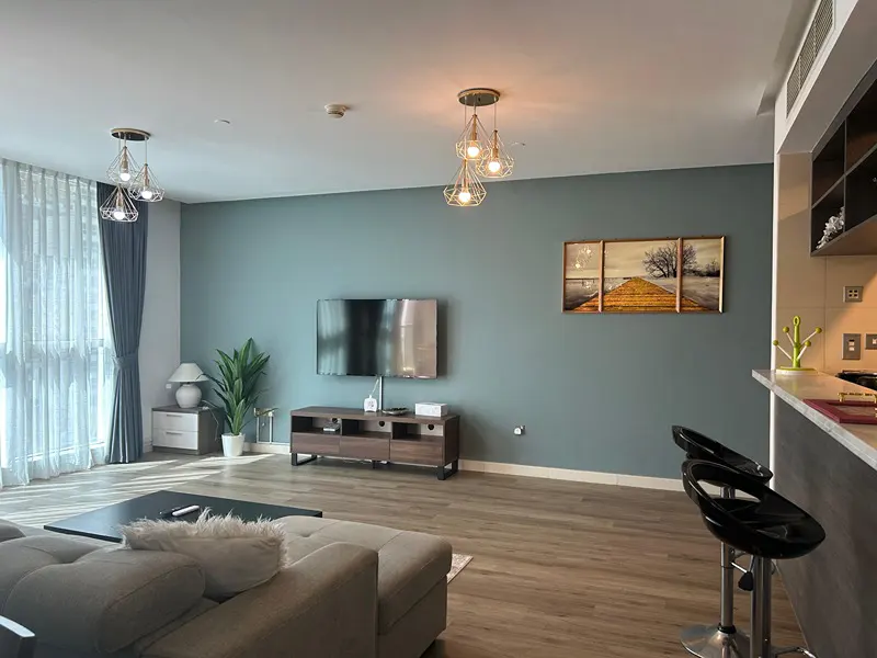 A modern living room with a gray sofa, TV, wooden floors, and teal walls. Three pendant lights hang from the ceiling.