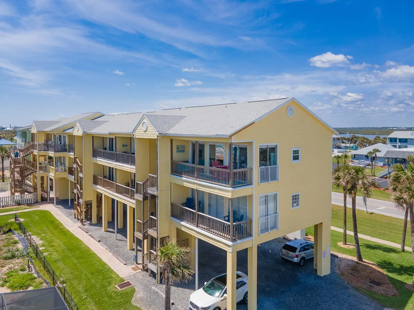 Aerial view of a yellow, three-story beach house with wooden balconies and stairs, elevated on stilts with cars parked underneath.