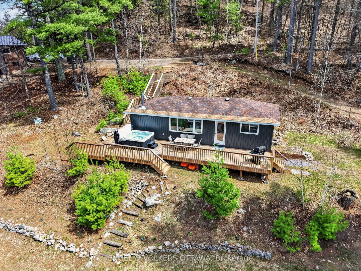 Aerial view of a gray house with a brown roof, a wooden deck with a hot tub, and a wooded background.