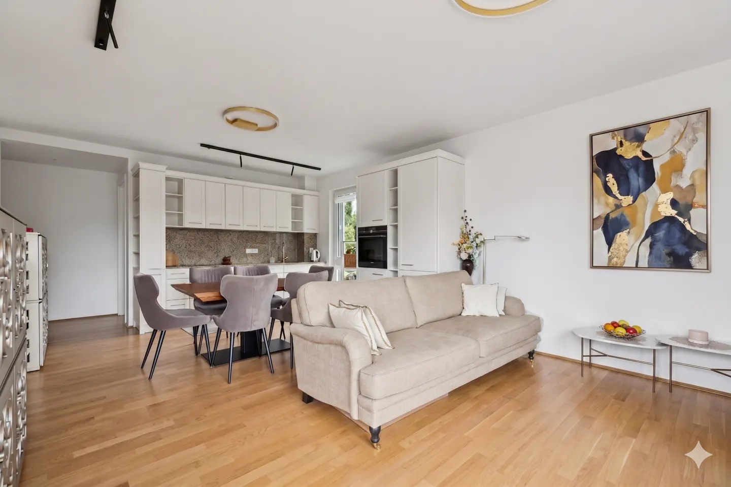 Bright, open-concept living space with a beige sofa, dining table, and white kitchen cabinets on a light wood floor. Abstract art on the wall.