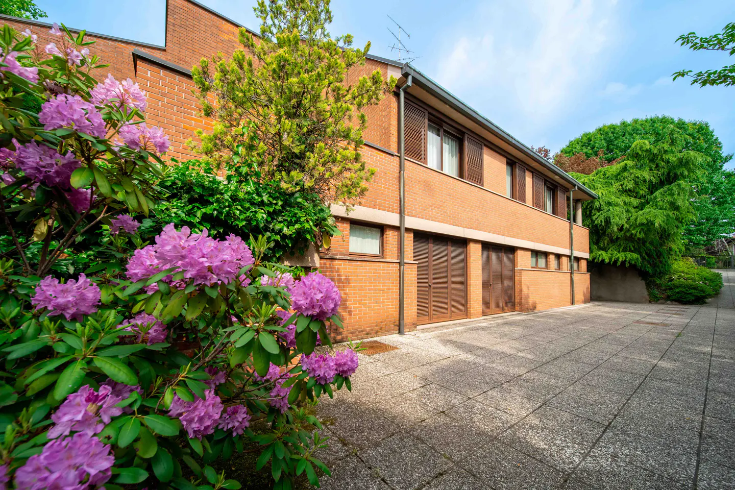 Brick building with brown shutters and garage doors, framed by blooming purple rhododendrons and green trees. Paved driveway.