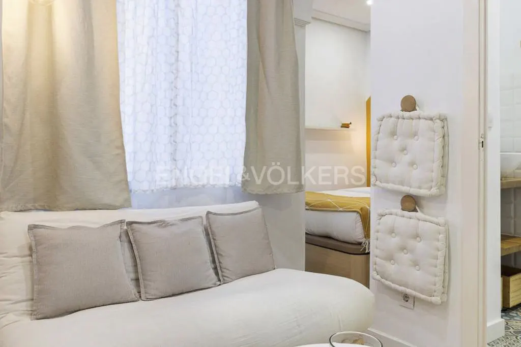 Living room with white sofa, beige pillows, and curtains. A bedroom and bathroom are visible in the background.