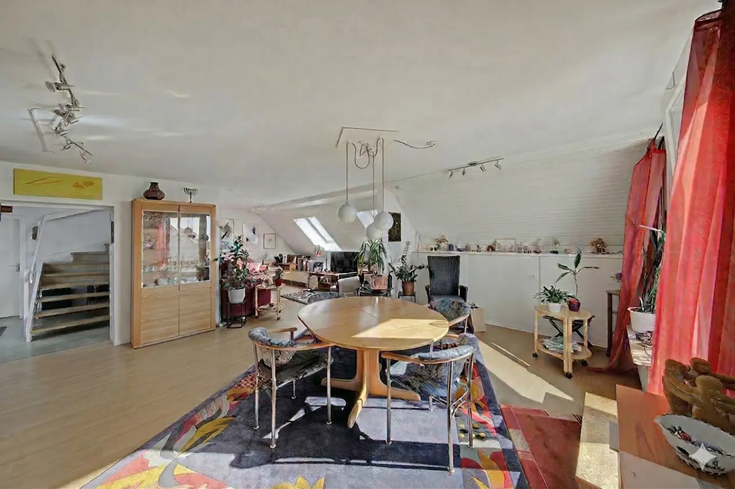 Attic living room with a round wooden table, four chairs, and a colorful rug. A staircase and display cabinet are visible in the background.