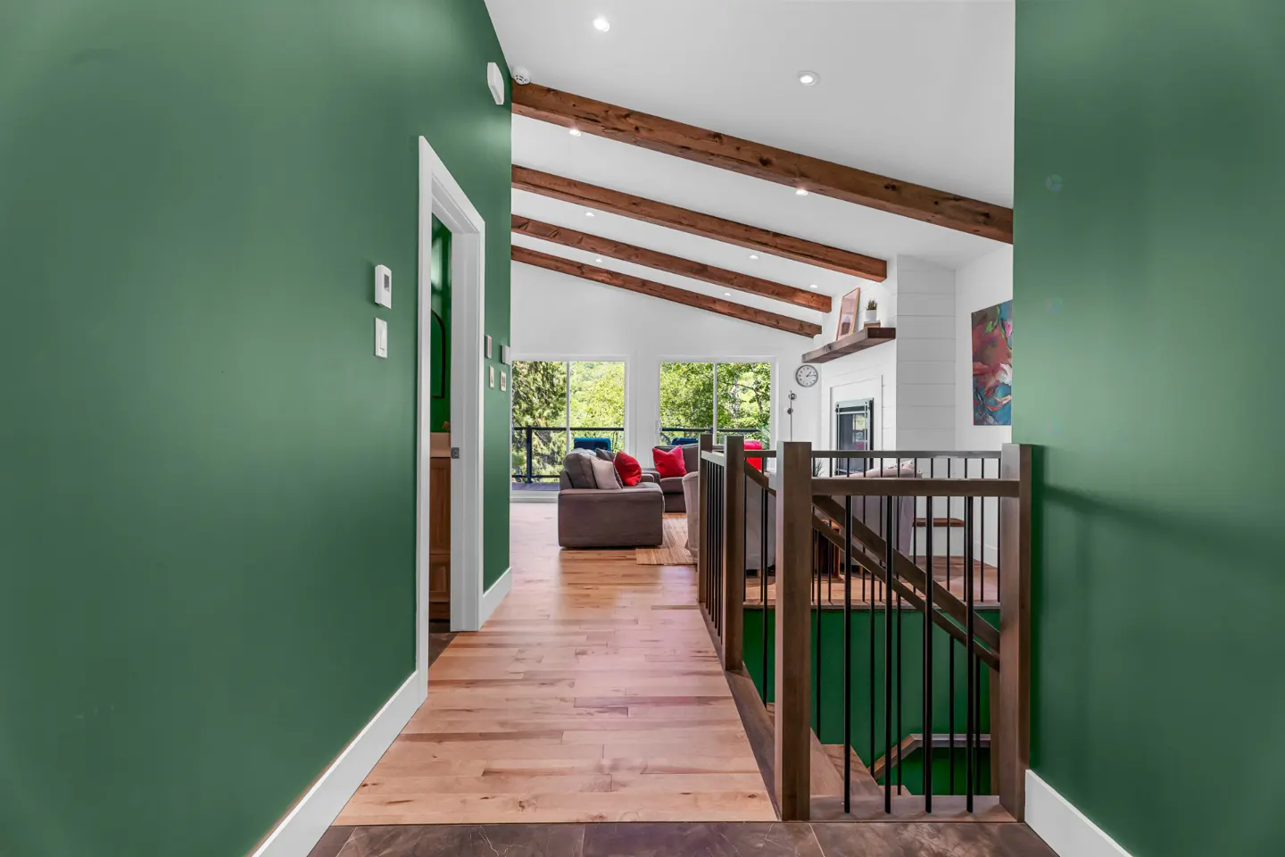 Hallway with green walls, wood floors, and a vaulted ceiling with exposed beams. Living room visible with sofas and large windows.
