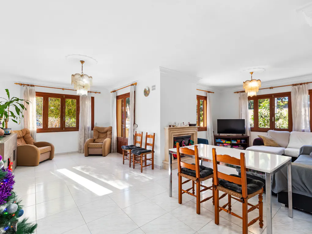 Bright living room with white tile floors, brown wood windows, and two tan armchairs. A dining table with four chairs sits in the center.