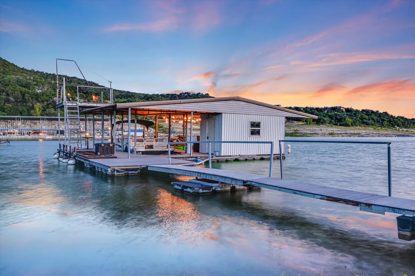 A boathouse on a lake at sunset. The boathouse has a white exterior and a metal roof. A dock leads to the boathouse. The sky is blue and orange.