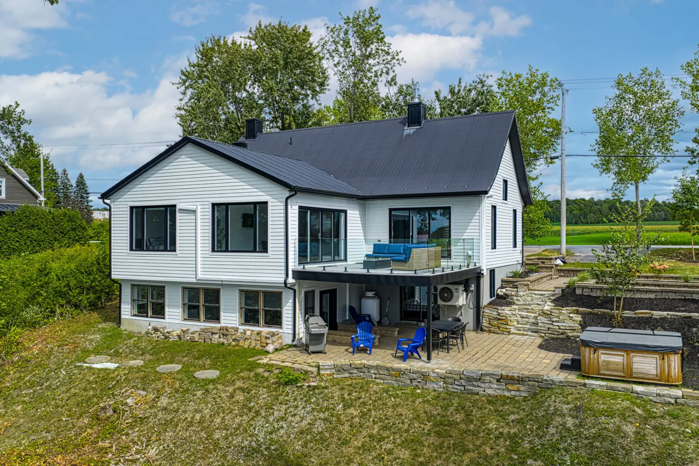 White two-story house with black roof, elevated deck, stone patio, hot tub, and blue outdoor furniture in a rural setting.
