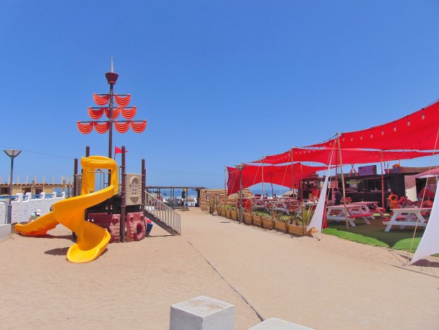 Beachside scene with a yellow playground, red canopies over picnic tables, and a clear blue sky.