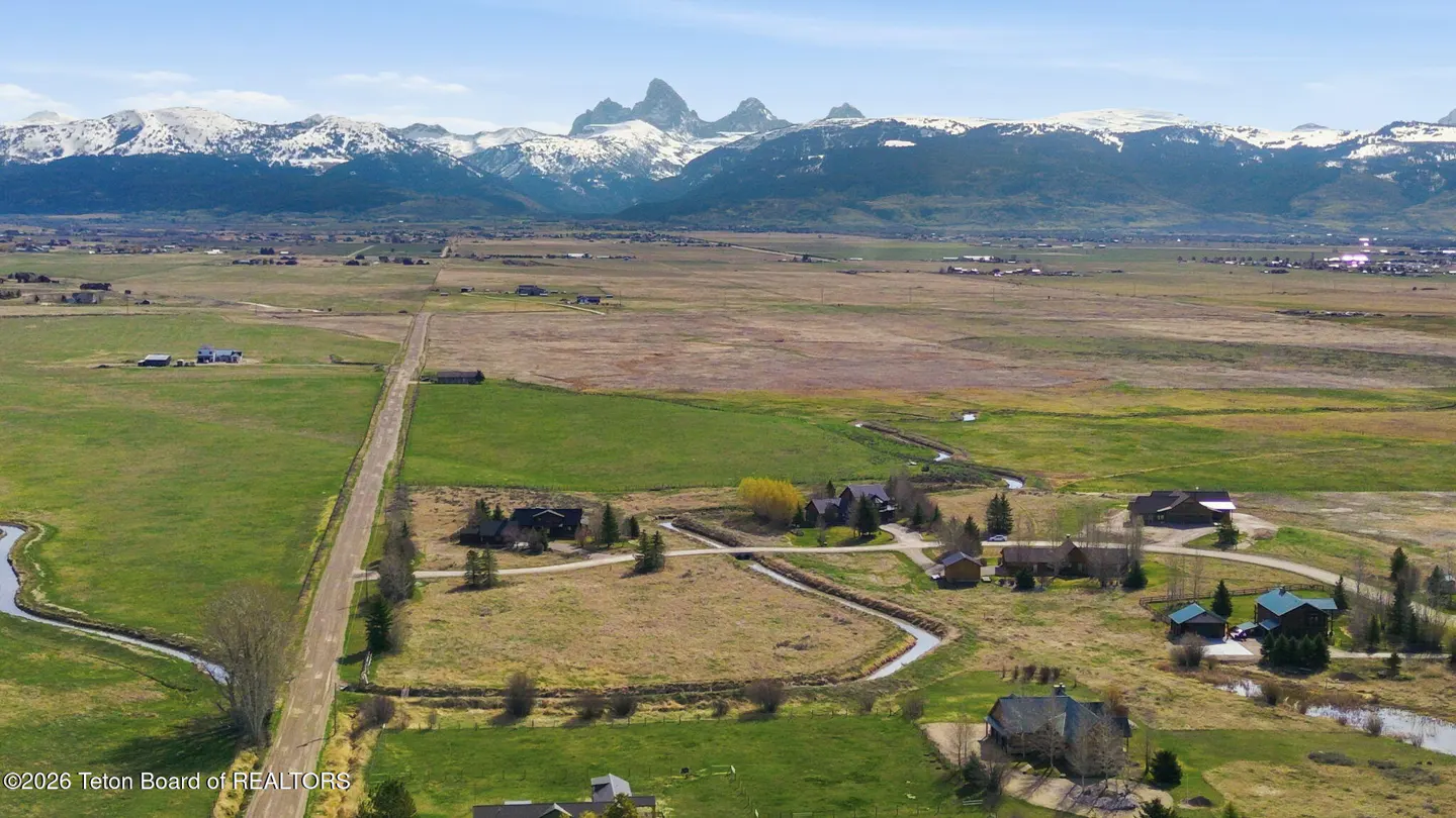Aerial view of a rural landscape with houses, fields, a road, and snow-capped mountains in the background.