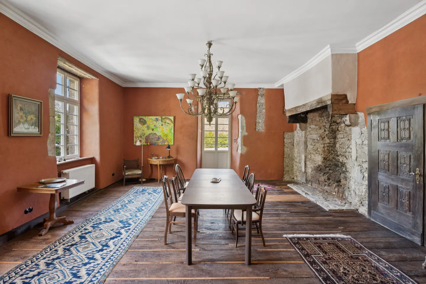 A dining room with orange walls, a long table, and a chandelier. A stone fireplace and a blue rug add character.