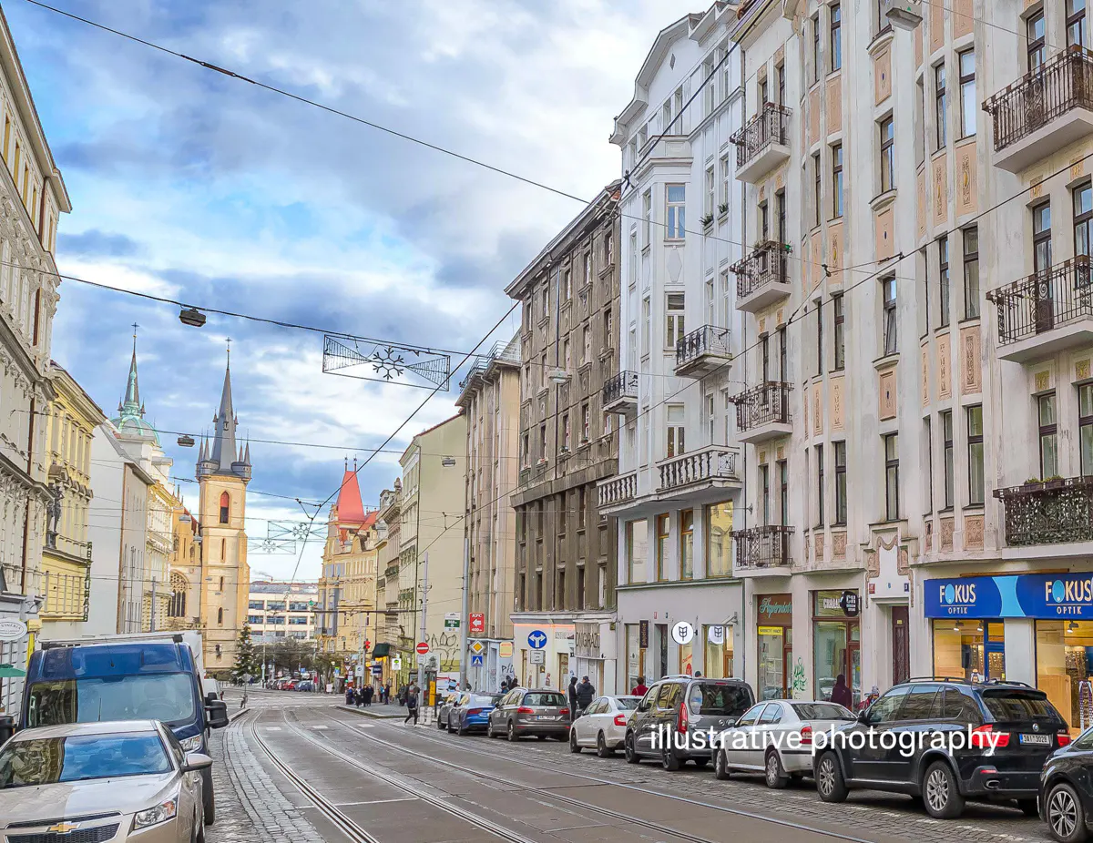 City street view with parked cars, tram tracks, and buildings. A church tower is visible in the distance under a cloudy sky.