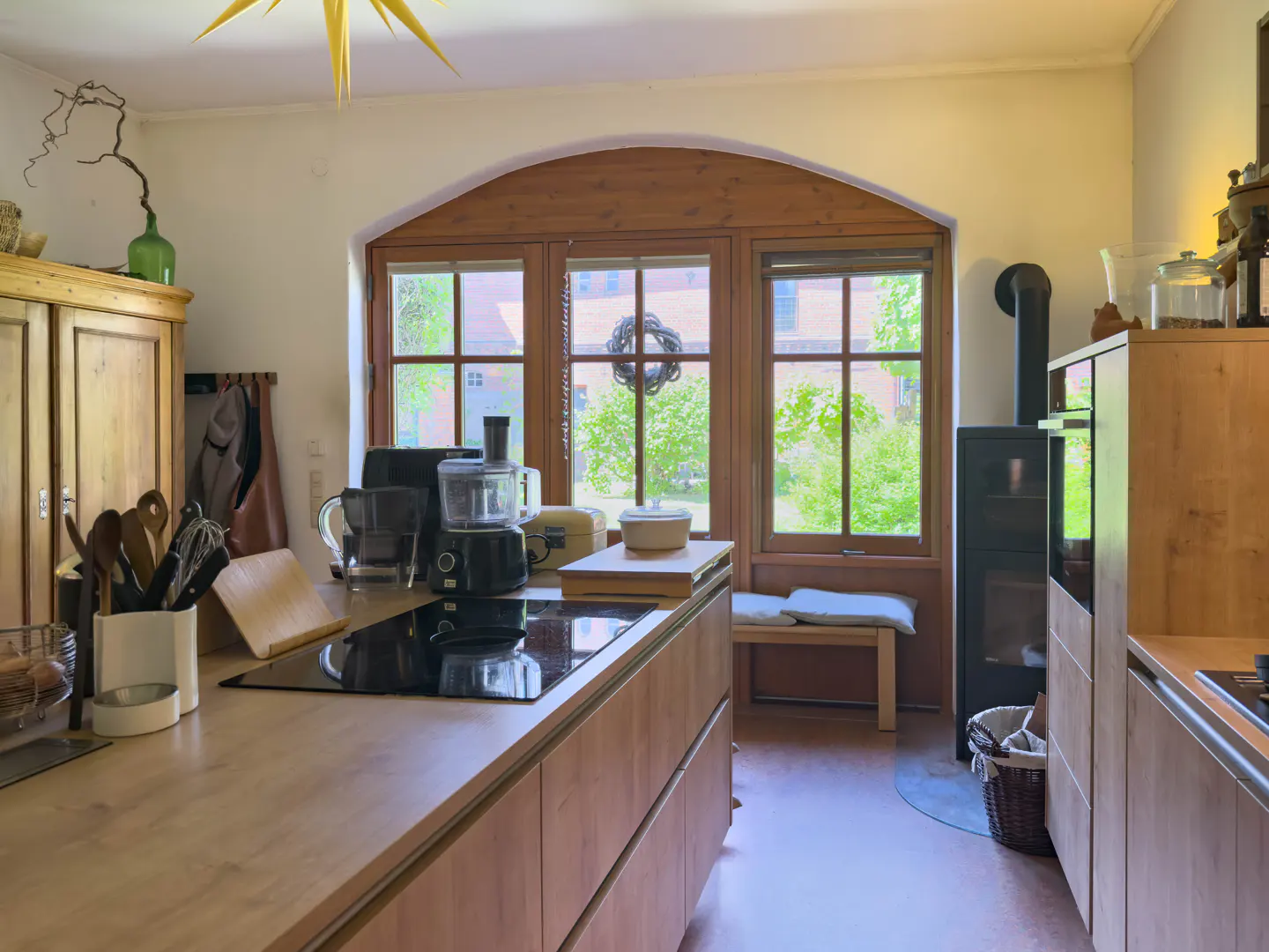 Bright kitchen with wood cabinets, black cooktop, and large arched window overlooking green trees. A wood-burning stove is visible.