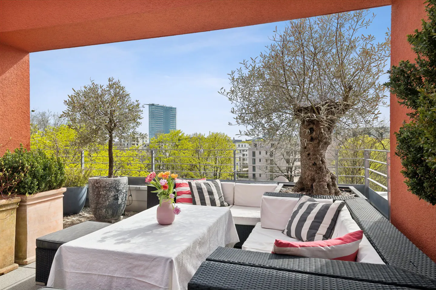 Outdoor patio with black wicker furniture, white cushions, and striped pillows. A white tablecloth covers the table with flowers in a pink vase. Trees and city views in the background.