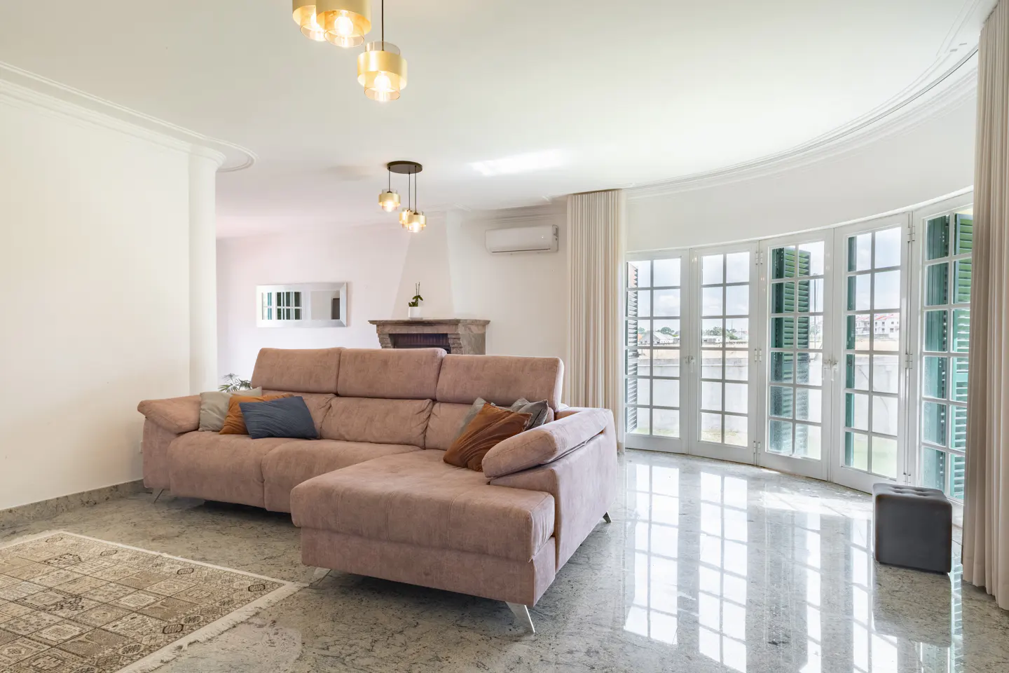 Bright living room with a pink sectional sofa, marble floors, and large windows with green shutters. Modern light fixtures hang from the white ceiling.