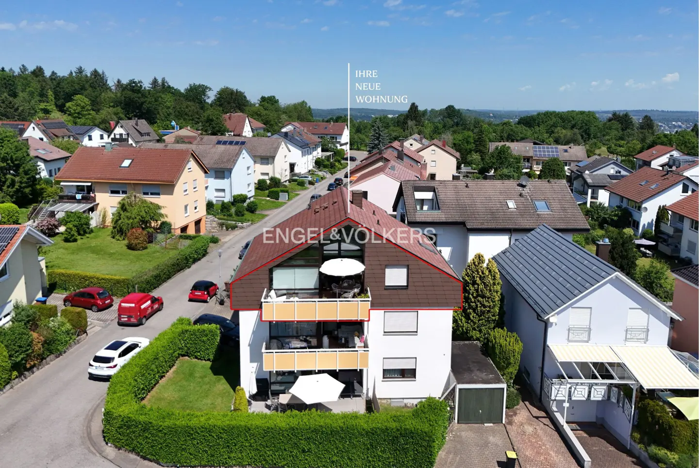 Aerial view of a two-story white house with brown roof and balconies, surrounded by other houses and greenery. "Engel & Völkers" logo visible.