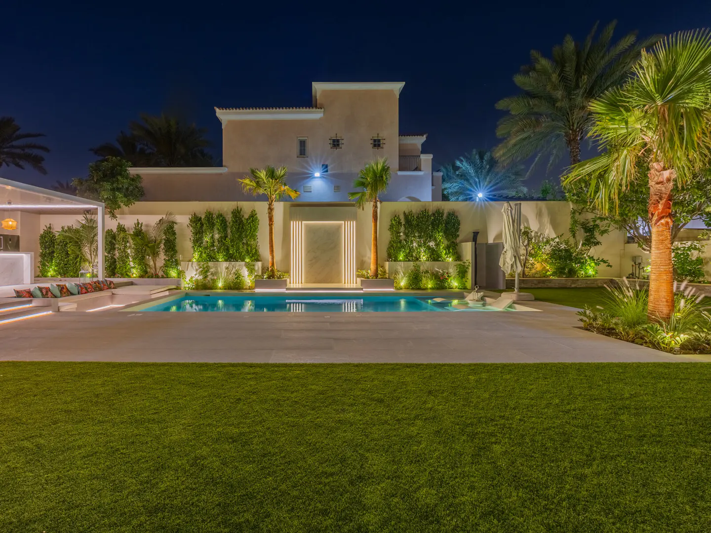 Night view of a luxury home's backyard with a pool, green lawn, palm trees, and a bar area with colorful pillows.