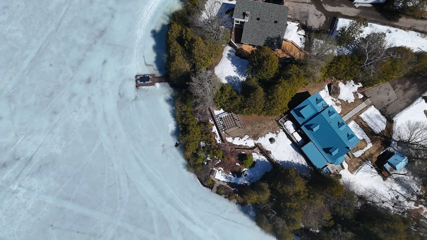 Aerial view of a lake with a frozen surface, a dock, and houses with green and blue roofs surrounded by trees and snow.