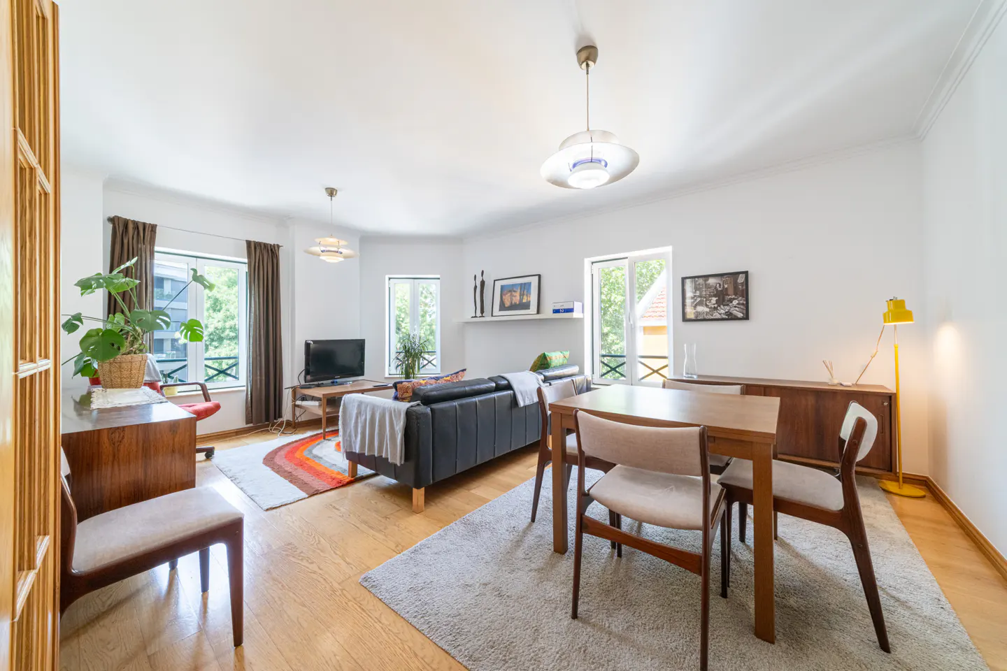 Bright living room with wood floors, white walls, and mid-century modern furniture. A gray sofa and dining table sit on a light gray rug.