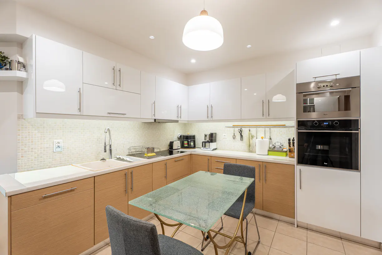 Bright kitchen with white cabinets, light wood lower cabinets, and a glass table with gray chairs. Stainless steel oven and microwave are built-in.